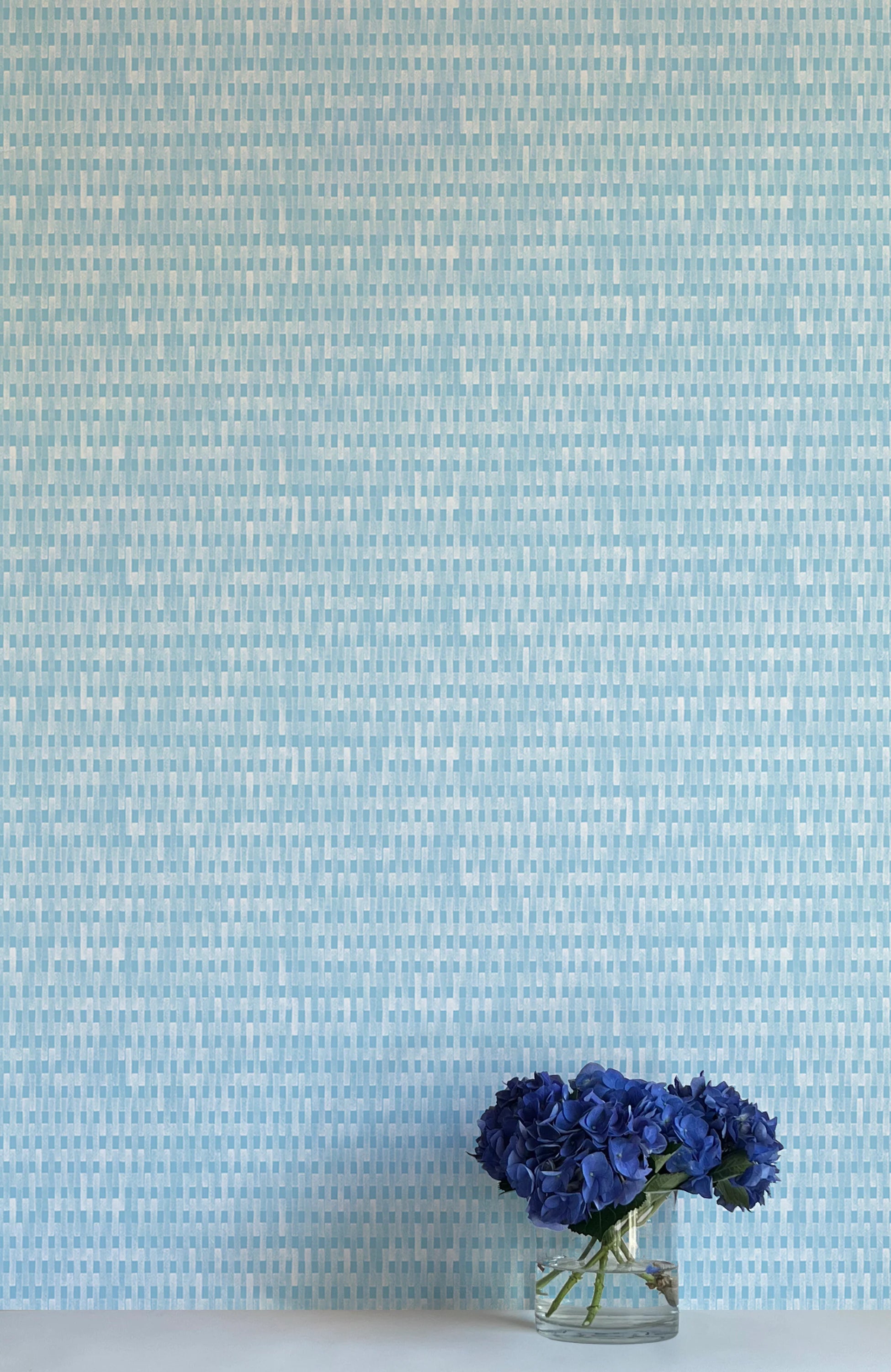 A vase of flowers stands in front of a wall covered in a textural grid print in shades of blue and cream.