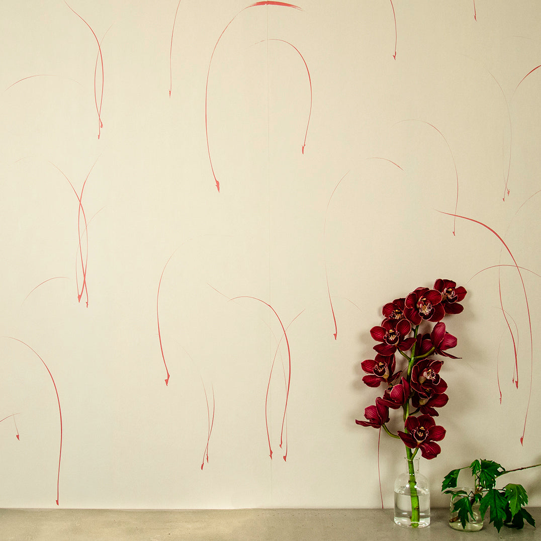 A vase of flowers stands in front of a wall papered in an elongated paint splatter pattern in red on a cream field.