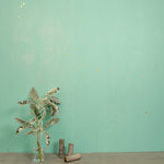 A table with a vase stands in front of a wall papered in a random splattered pattern in metallic gold on a green field.