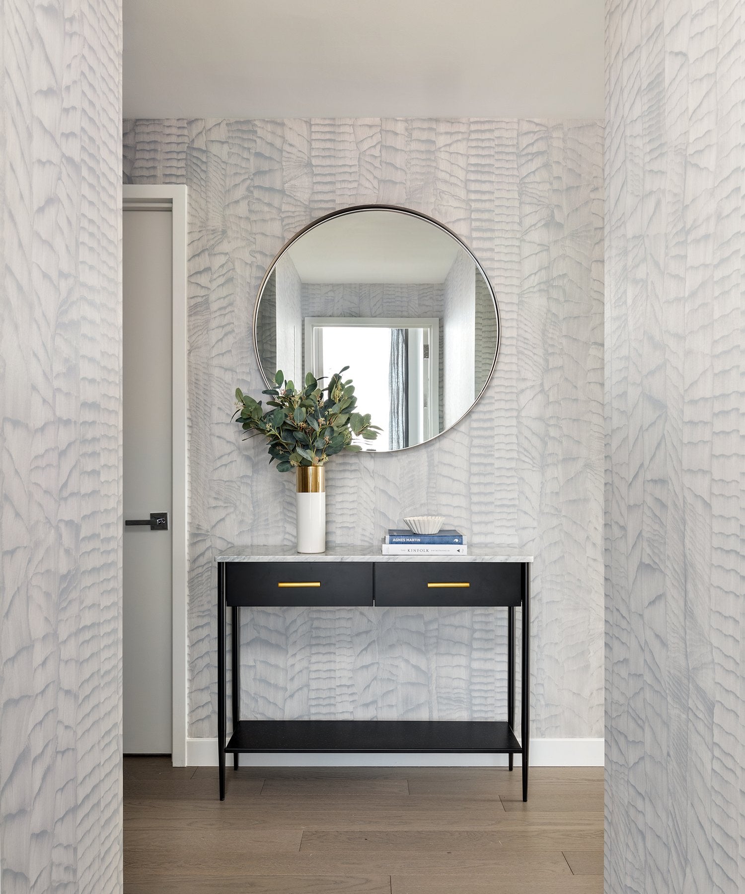 A hallway with a modernist end table and mirror and walls papered in an abstract textural pattern in metallic gray.