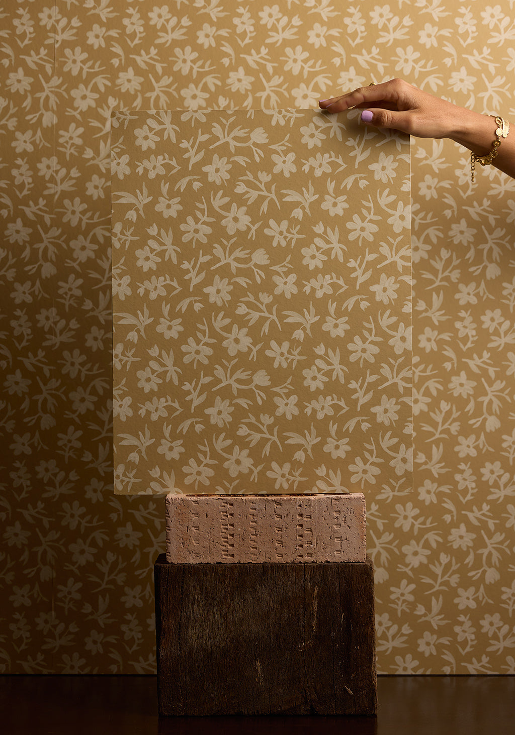 Hand holding a sheet of floral-patterned wallpaper against a matching wall.