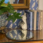 An end table with books, a plant and a mirrored plate stands in front of a wall papered in a falling leaf print.