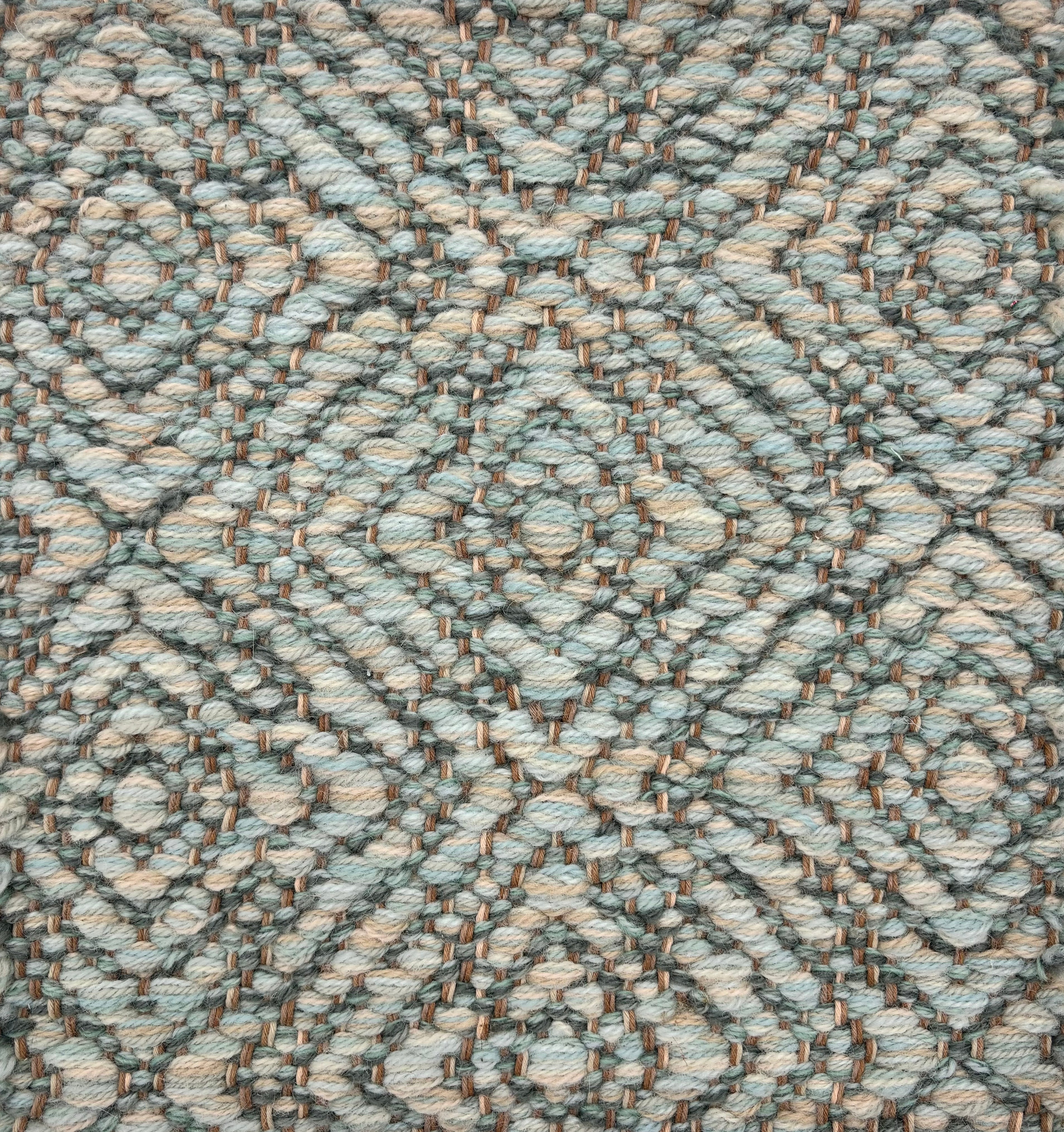 Handwoven rug sample in a radiating diamond pattern in shades of soft aqua, cream and green.