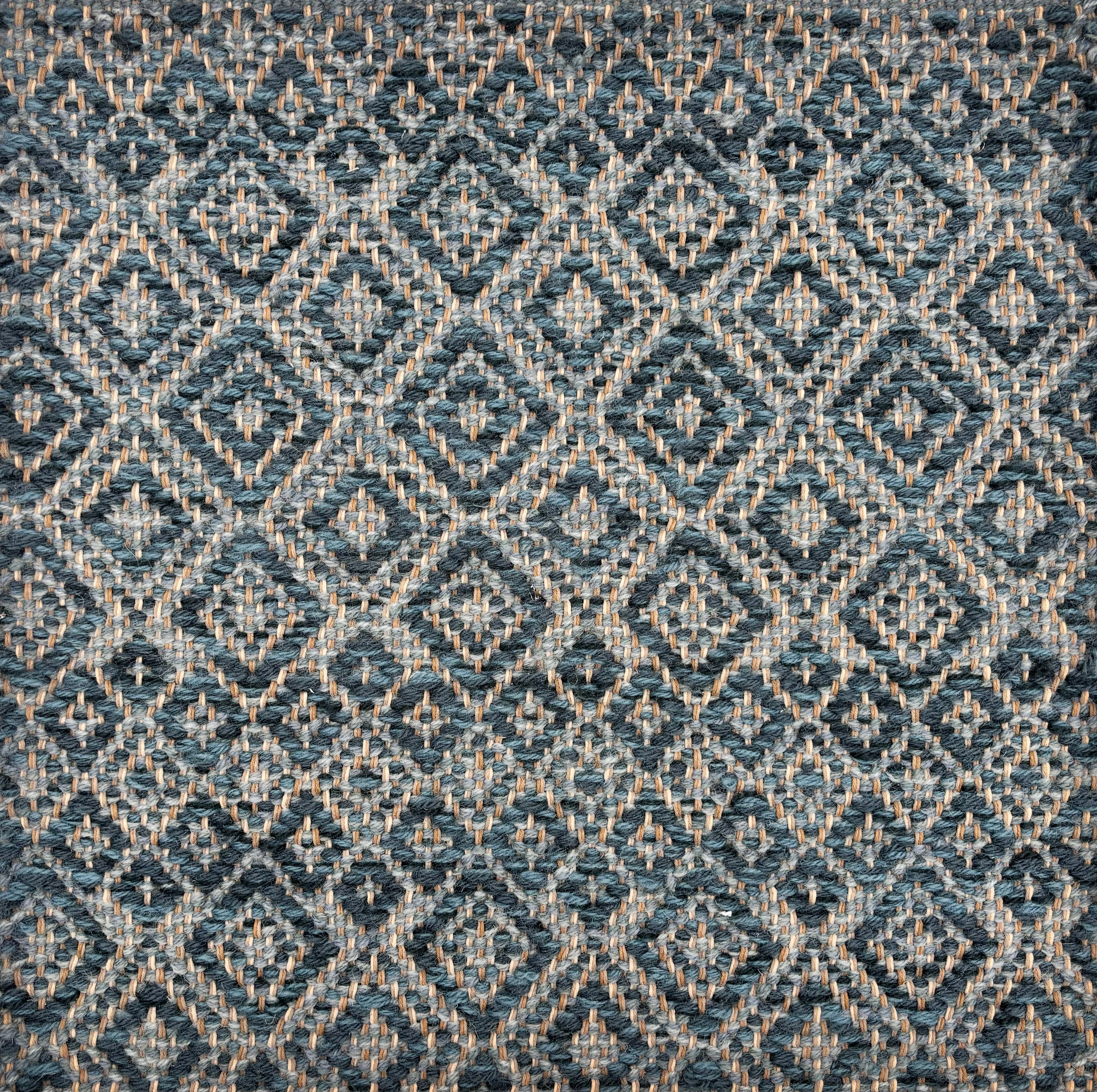 Handwoven rug sample in a radiating diamond pattern in shades of teal blue, grey, and tan. 