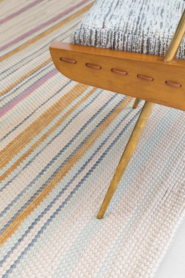 A wooden chair stands on a woven leather rug in an irregular stripe print in shades of pink, yellow and blue.