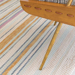 A wooden chair stands on a woven leather rug in an irregular stripe print in shades of pink, yellow and blue.