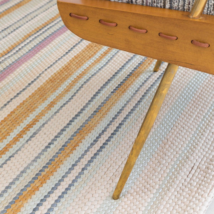 A wooden chair stands on a woven leather rug in an irregular stripe print in shades of pink, yellow and blue.