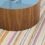 A round coffee table stands on a woven leather rug in an irregular stripe print in shades of pink, yellow and blue.