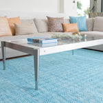 A glass coffee table stands on a woven leather rug in a striped wave texture in shades of turquoise with cream accents.