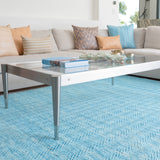 A glass coffee table stands on a woven leather rug in a striped wave texture in shades of turquoise with cream accents.