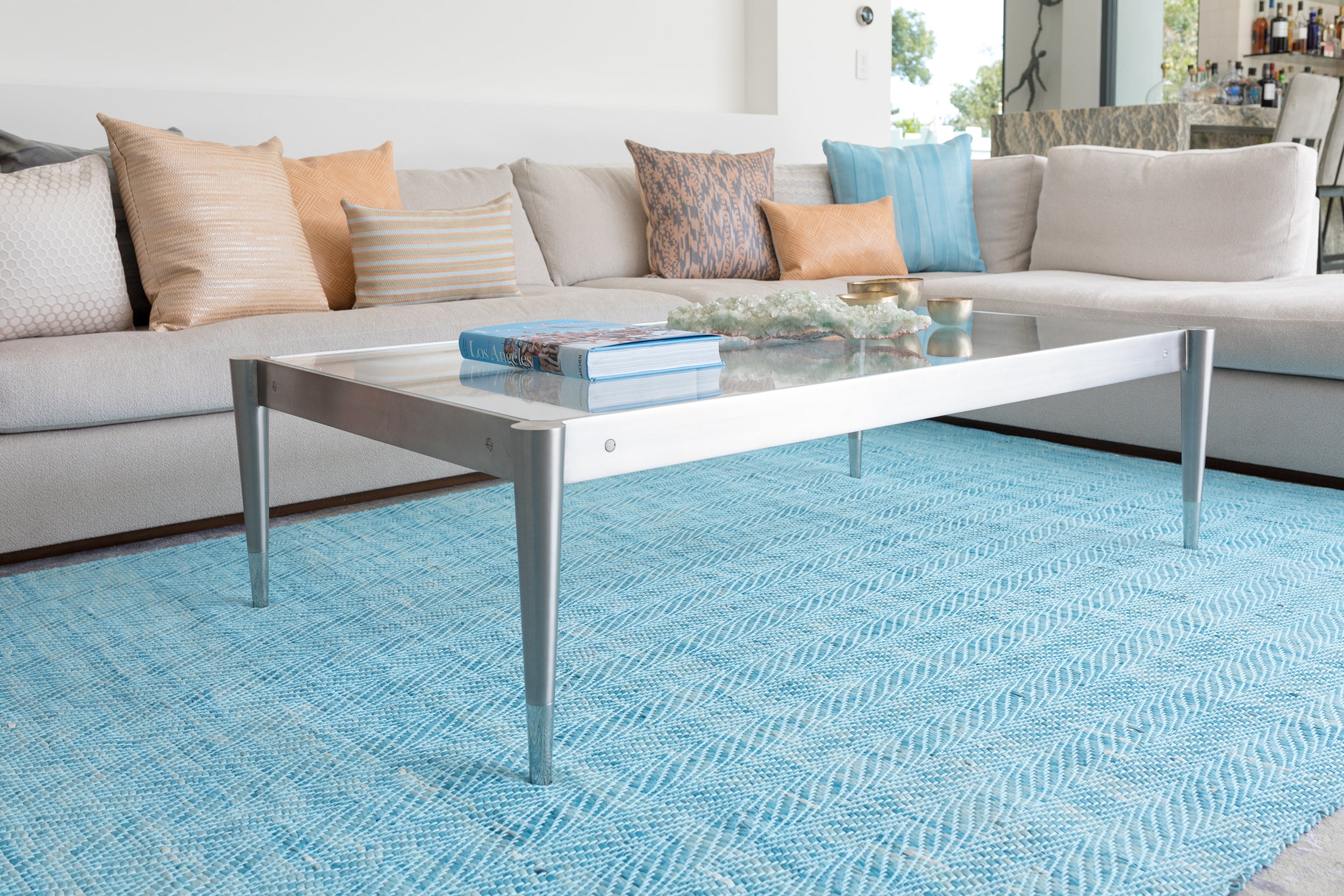 A glass coffee table stands on a woven leather rug in a striped wave texture in shades of turquoise with cream accents.