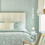 A bedroom tableau with a headboard and bedding upholstered in patterned textiles in shades of light turquoise, white and yellow.