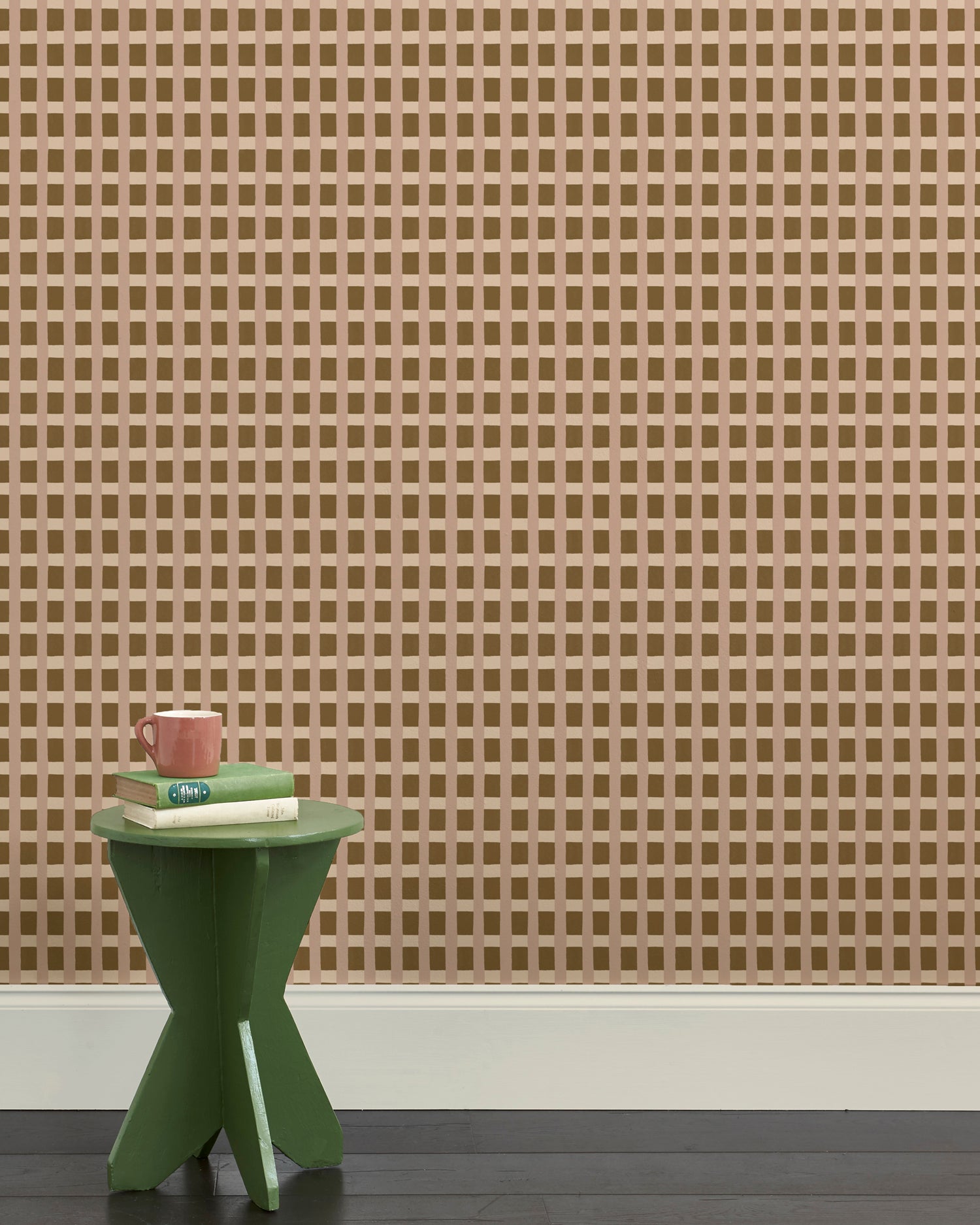 A green wooden stool with books and a mug against a wallpapered wall in a gridded check pattern in brown, cream and mauve. 