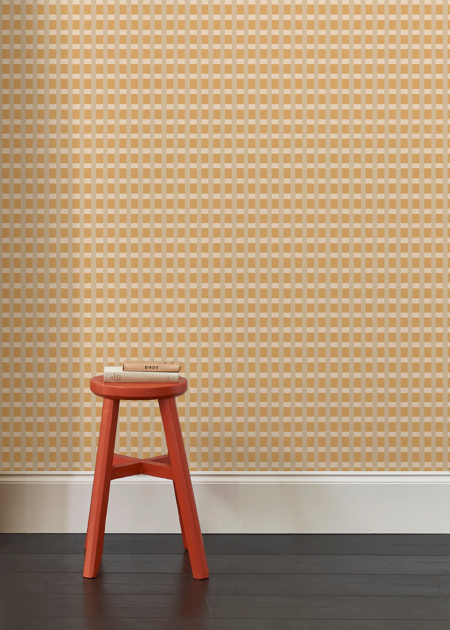 A green red stool with books  a wallpapered wall in a gridded check pattern in tan, cream ochre yellow. 