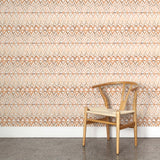 A wooden chair stands in front of a wall papered in a painterly ikat print in shades of tan and orange on a white field.