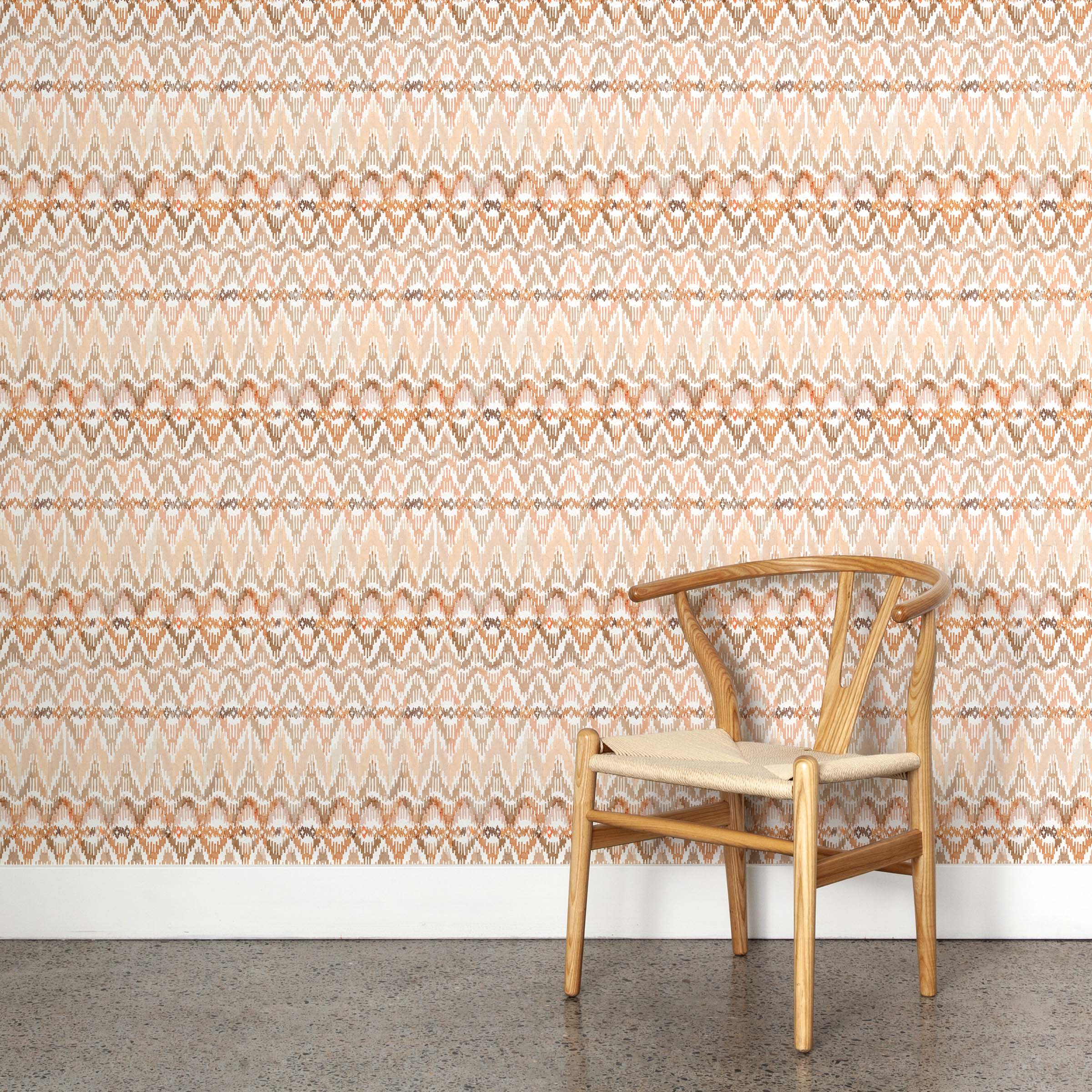 A wooden chair stands in front of a wall papered in a painterly ikat print in shades of tan and orange on a white field.