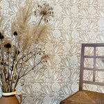 A chair and bunch of flowers stands in front of a wall papered in a dense floral print in white on a tan field.