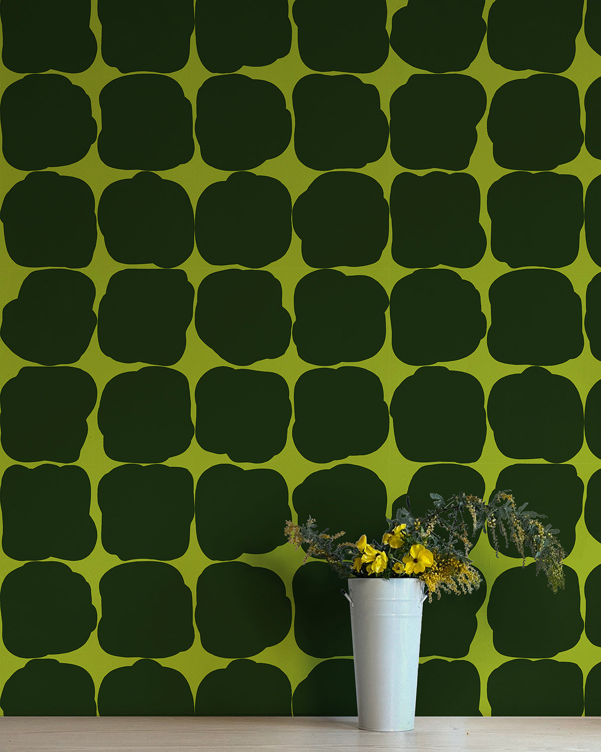 A vase of flowers stands in front of a wall papered in a repeating blot print in black on an olive field.