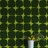 A vase of flowers stands in front of a wall papered in a repeating blot print in black on an olive field.