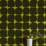 A vase of flowers in front of a wall papered in a repeating blot print in dark green on a green field.