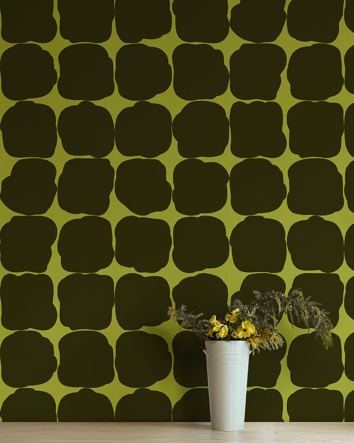 A vase of flowers in front of a wall papered in a repeating blot print in dark green on a green field.