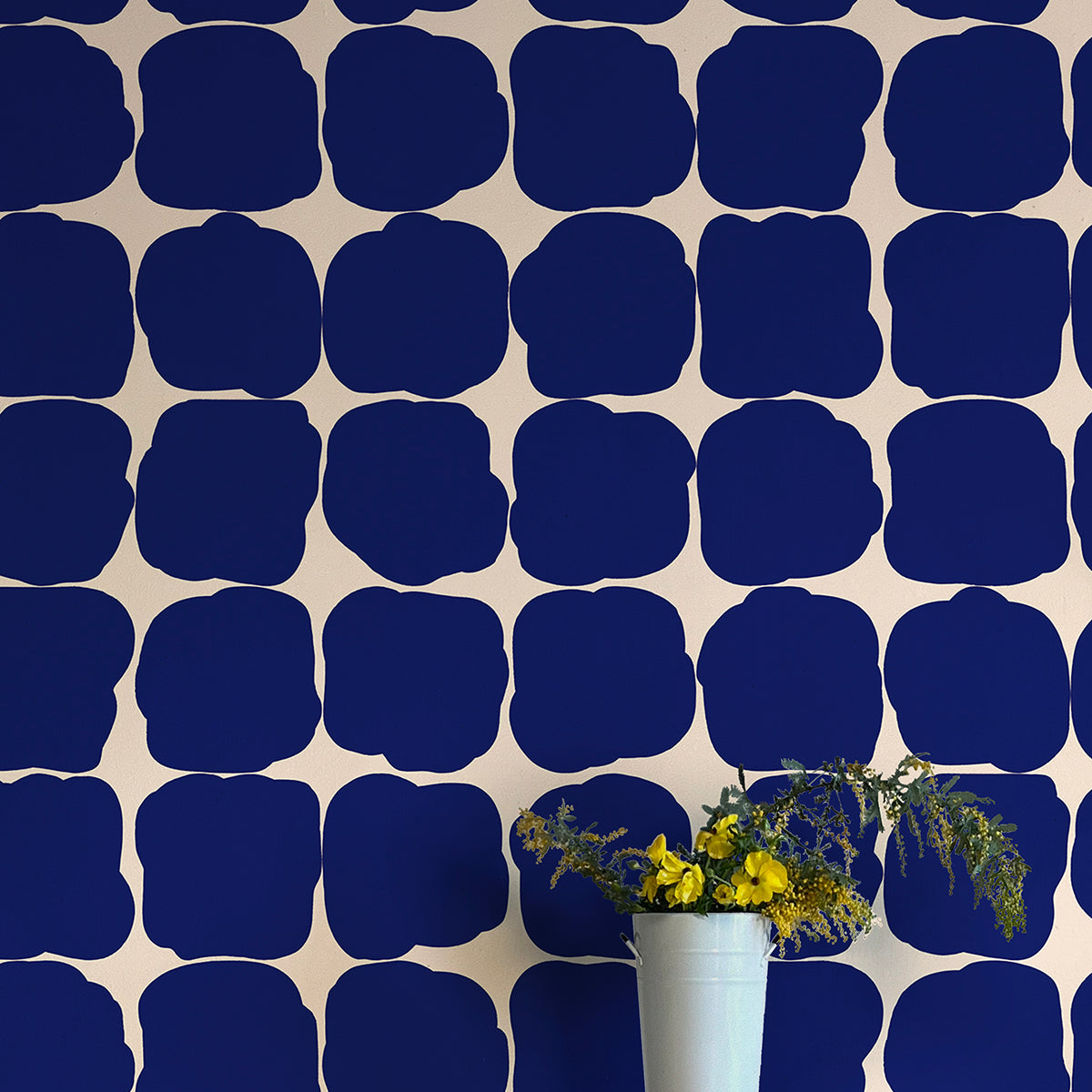 A vase of flowers stands in front of a wall papered in a repeating blot print in navy on a cream field.