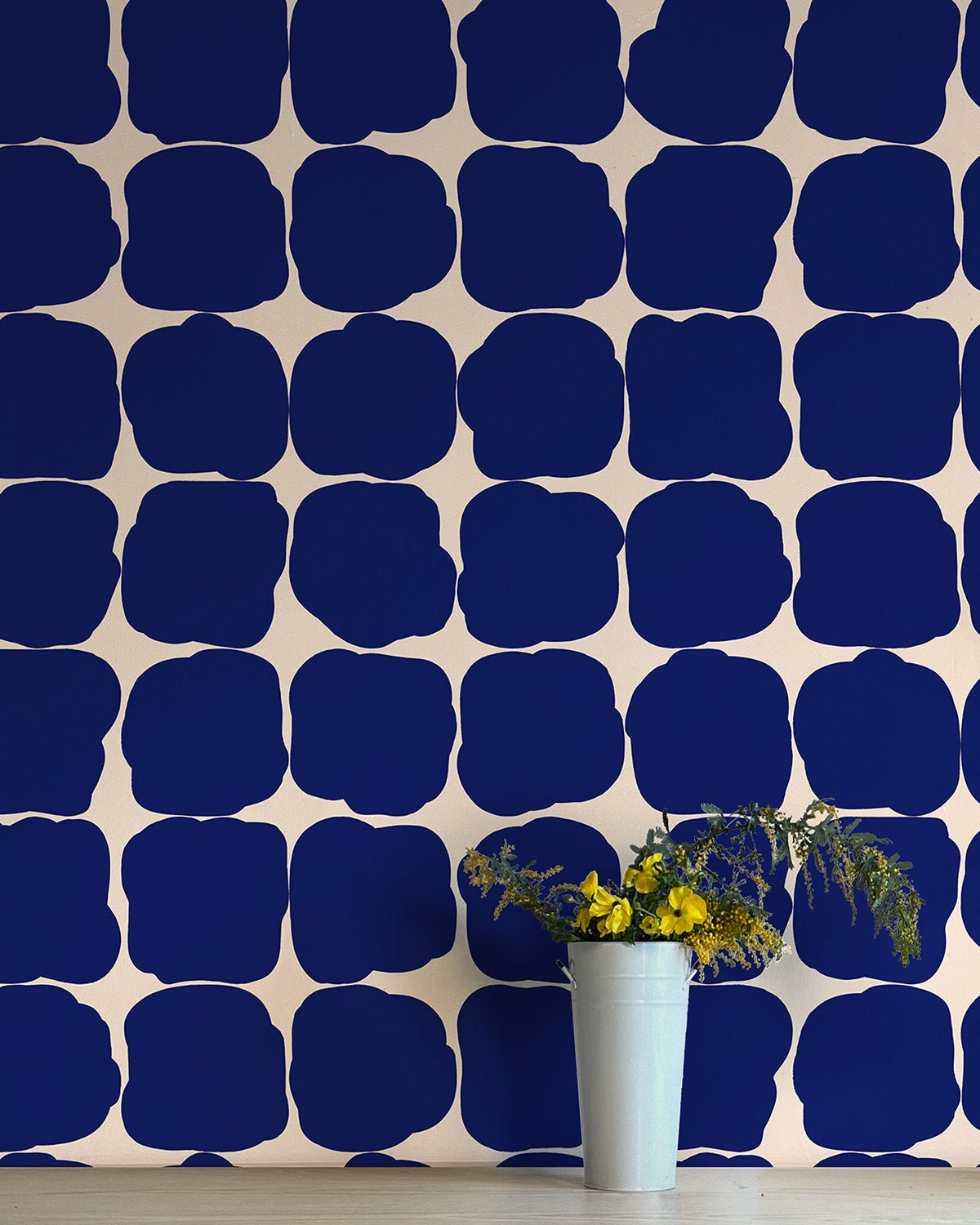 A vase of flowers stands in front of a wall papered in a repeating blot print in navy on a cream field.