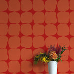 A vase of flowers stands in front of a wall papered in a repeating blot print in orange on a red field.