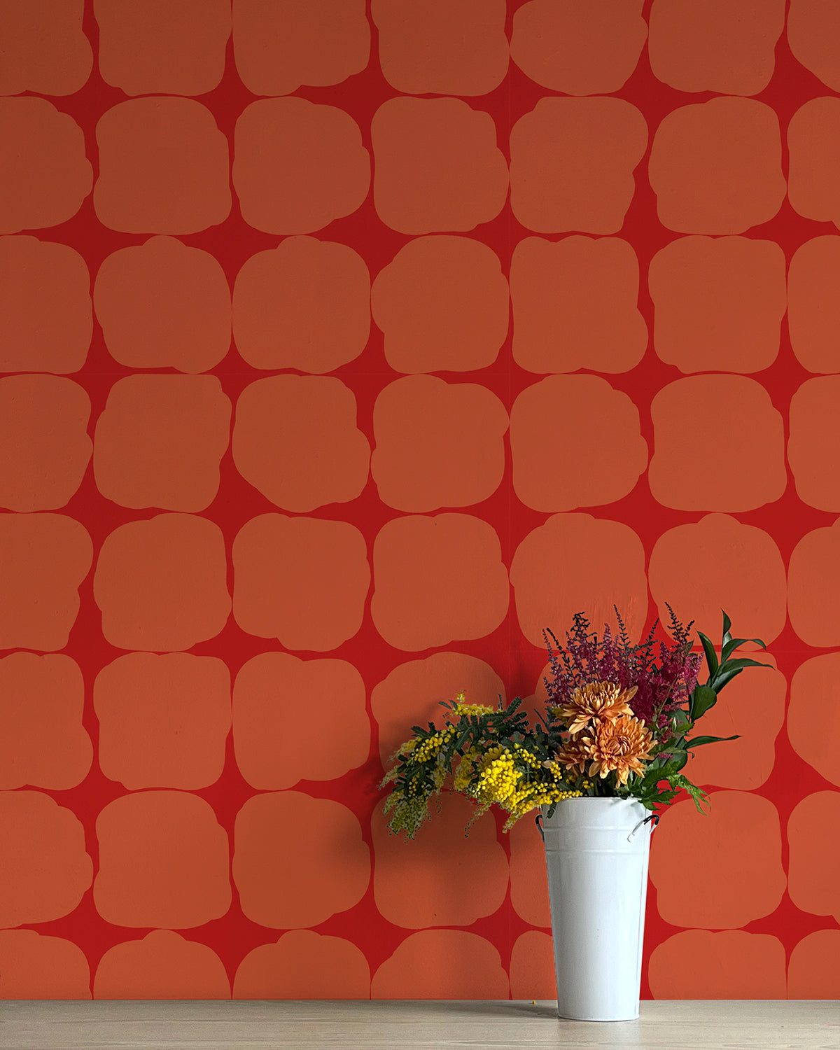 A vase of flowers stands in front of a wall papered in a repeating blot print in orange on a red field.