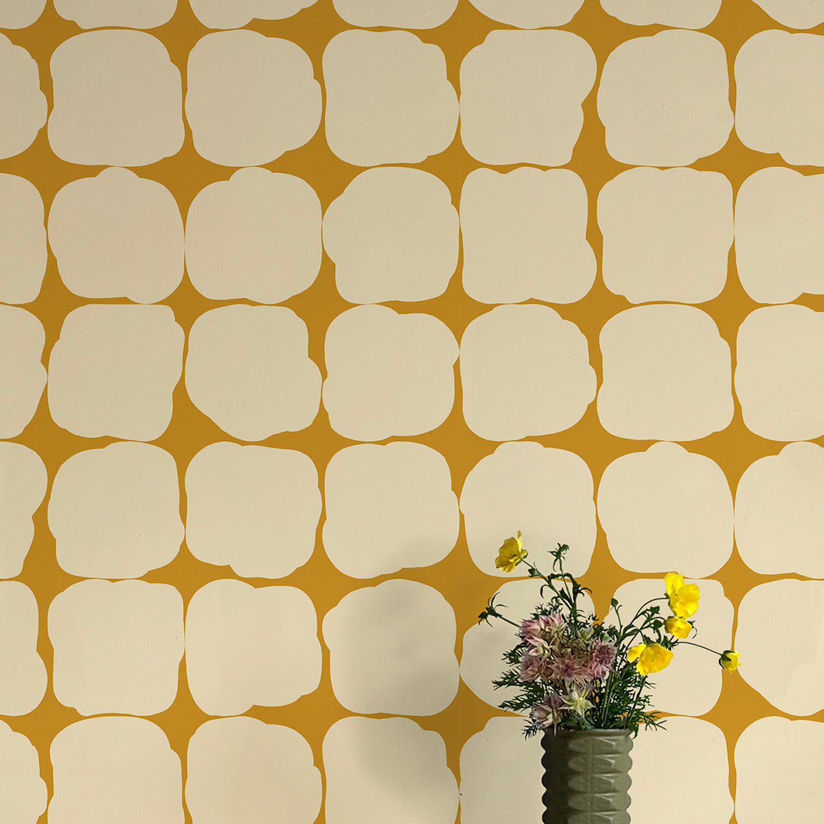 A vase of flowers stands in front of a wall papered in a repeating blot print in cream on a yellow field.