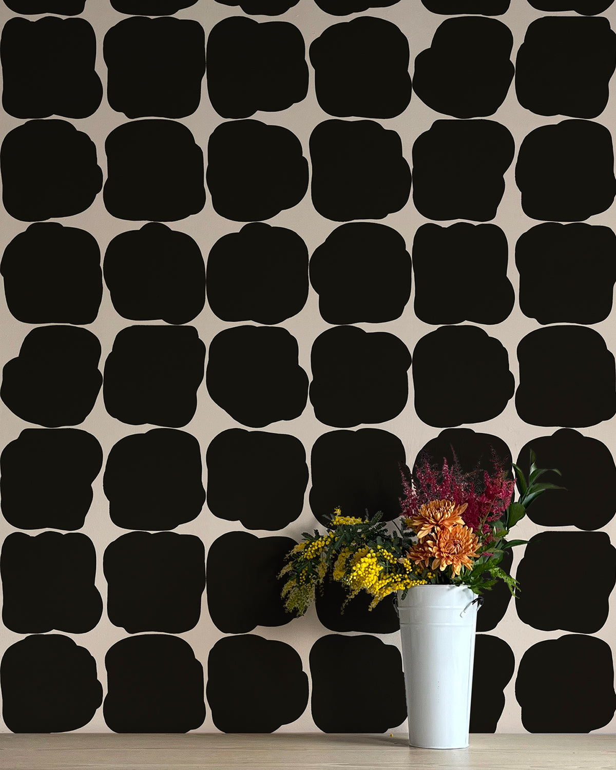 A vase of flowers stands in front of a wall papered in a repeating blot print in black on a white field.