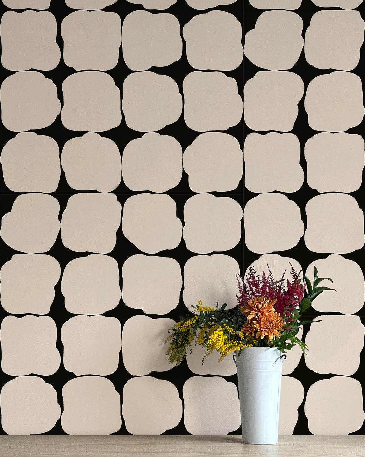 A vase of flowers stands in front of a wall papered in a repeating blot print in white on a black field.