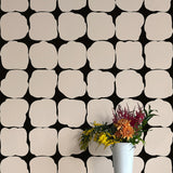 A vase of flowers stands in front of a wall papered in a repeating blot print in white on a black field.