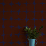 A vase of flowers stands in front of a wall papered in a repeating blot print in deep red on a dark blue field.