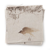 A stack of linen fabric swatches in a painterly bird and branch print in brown on a tan field.