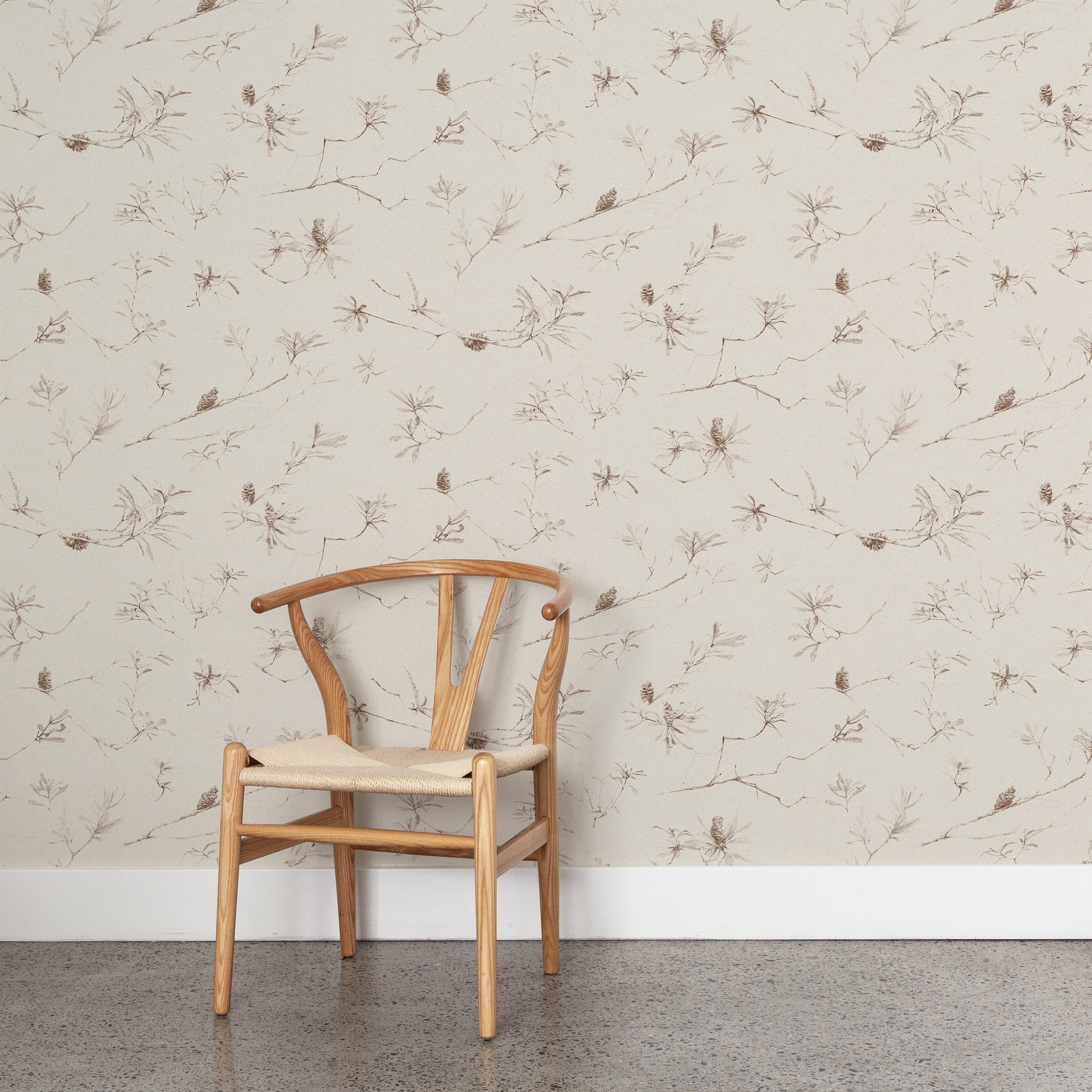 A wooden chair stands in front of a wall papered in a painterly pine cone and branch print in brown on a cream field.