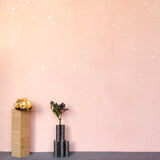 A table with knick knacks stands in front of a wall papered in a random splattered pattern in metallic cream on a pink field.