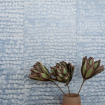 A vase of flowers stand in front of a wall papered in a repeating dappled texture in light blue.