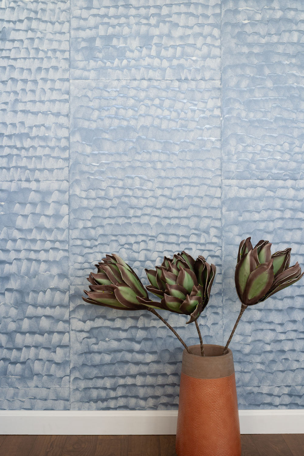 A vase of flowers stand in front of a wall papered in a repeating dappled texture in light blue.