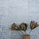 A vase of flowers stand in front of a wall papered in a repeating dappled texture in light blue.