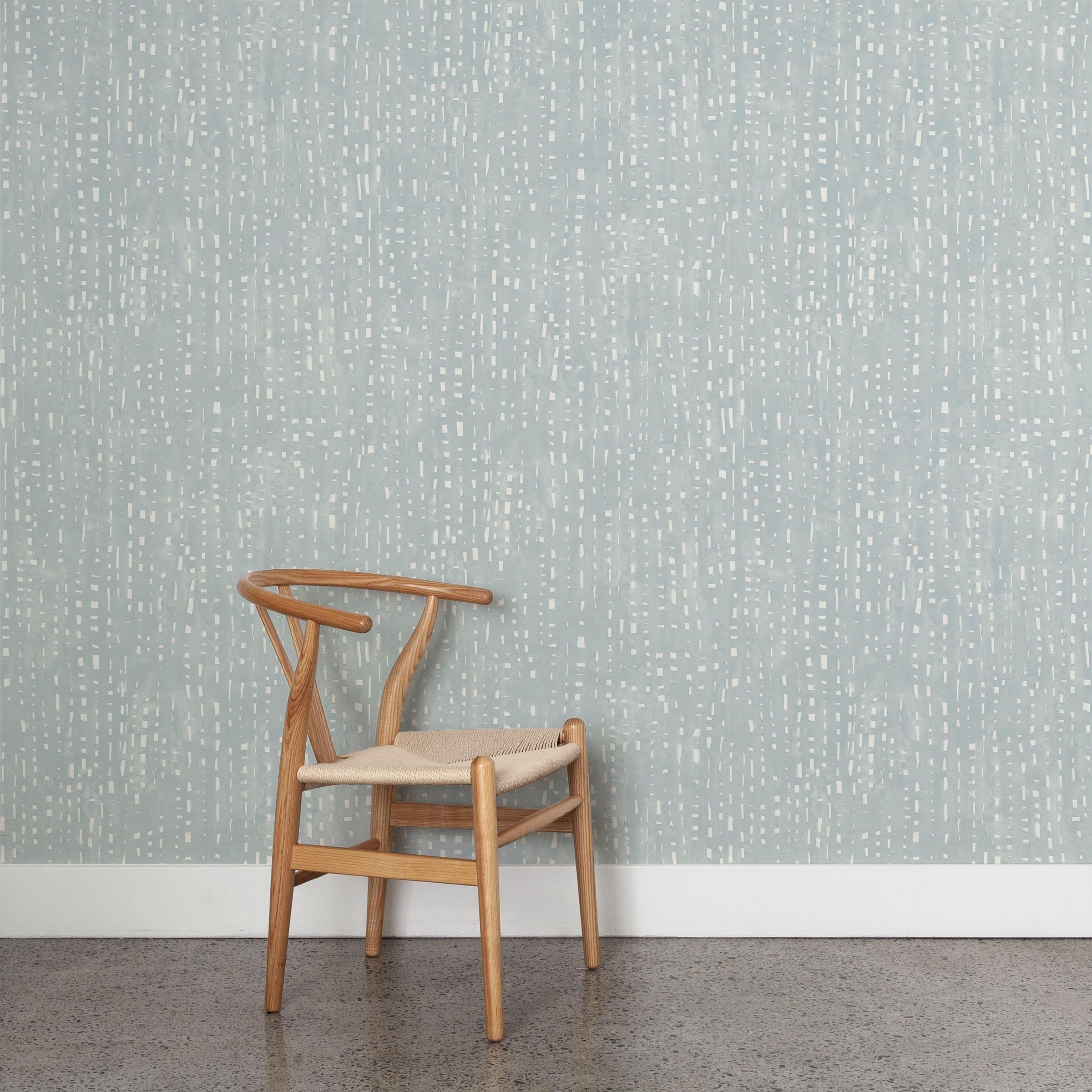 A wooden chair stands in front of a wall papered in a painterly small-scale grid print in blue-gray on a white field.