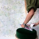 A woman opens a catalog in front of a wall papered in an abstract painted print in shades of green, yellow and white.