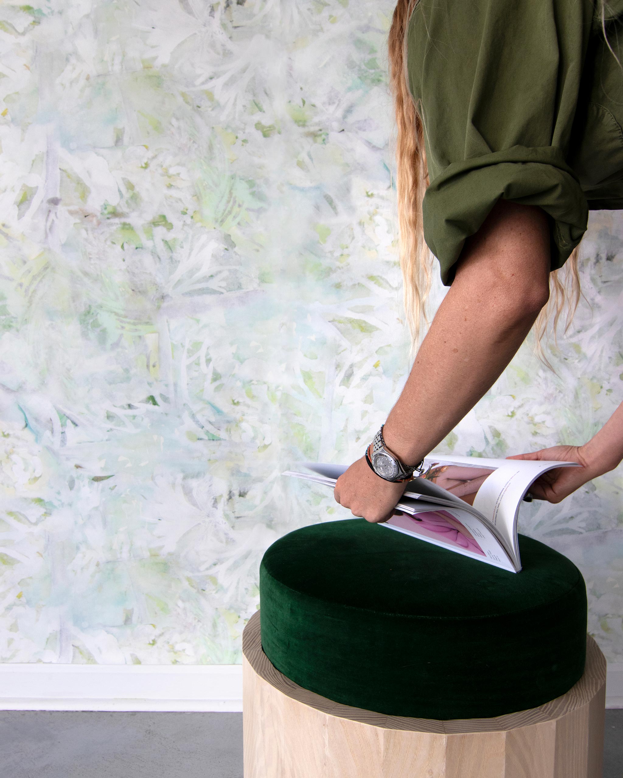 A woman opens a catalog in front of a wall papered in an abstract painted print in shades of green, yellow and white.