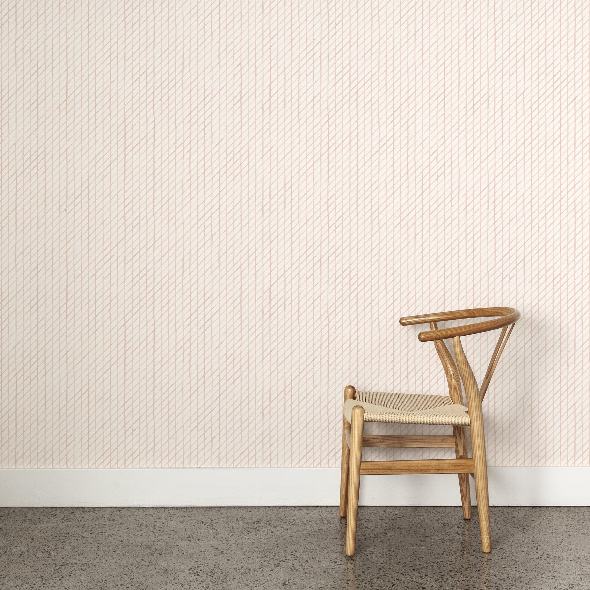 A wooden chair stands in front of a wall papered in a painterly uneven grid pattern in light pink on a white field.