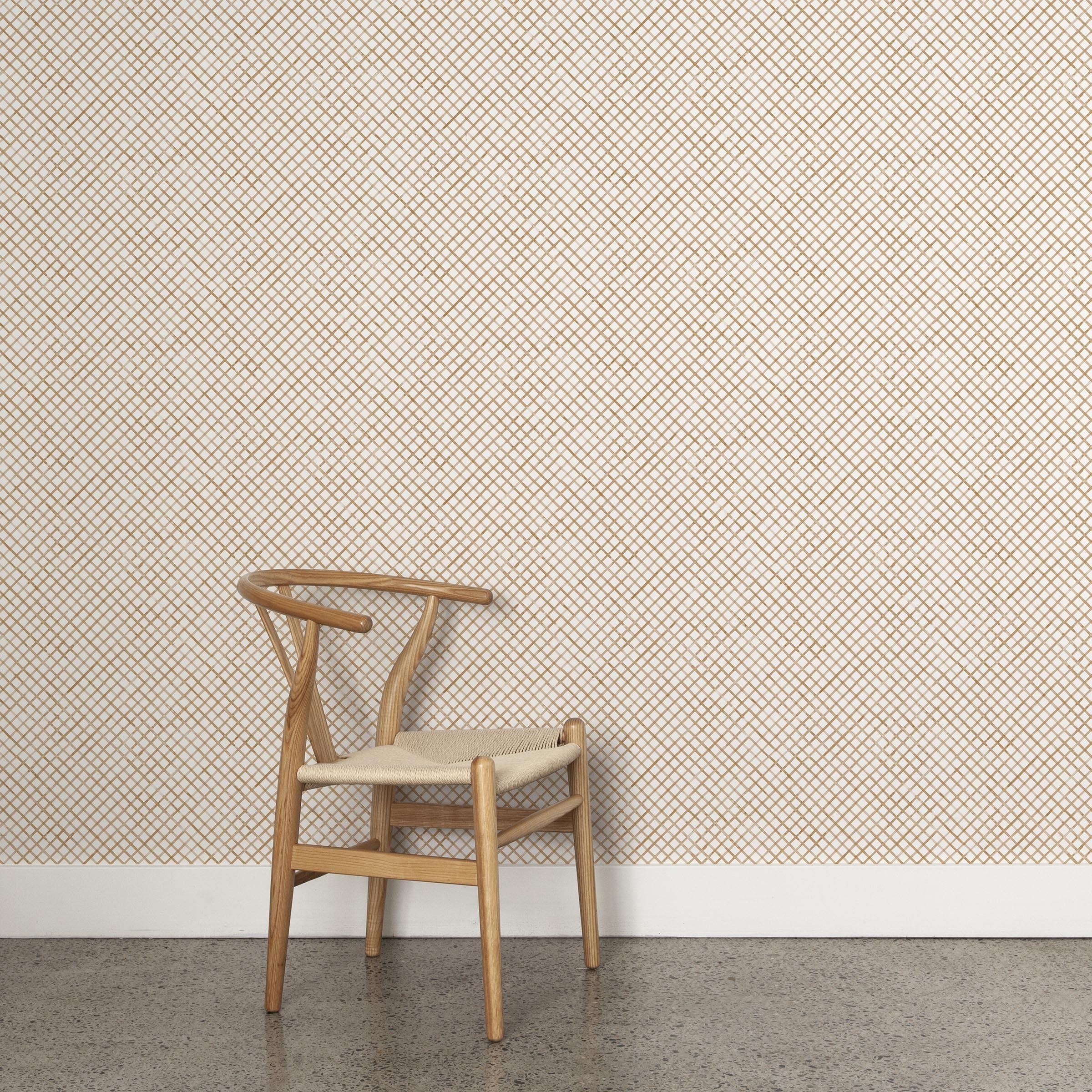 A wooden chair stands in front of a wall papered in a painterly grid pattern in bronze on a white field.