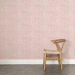 A wooden chair stands in front of a wall papered in an intricate striped grid pattern in dusty rose on a white field.