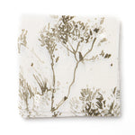 A stack of fabric swatches in a painterly shrub and tree print in olive on a cream field.