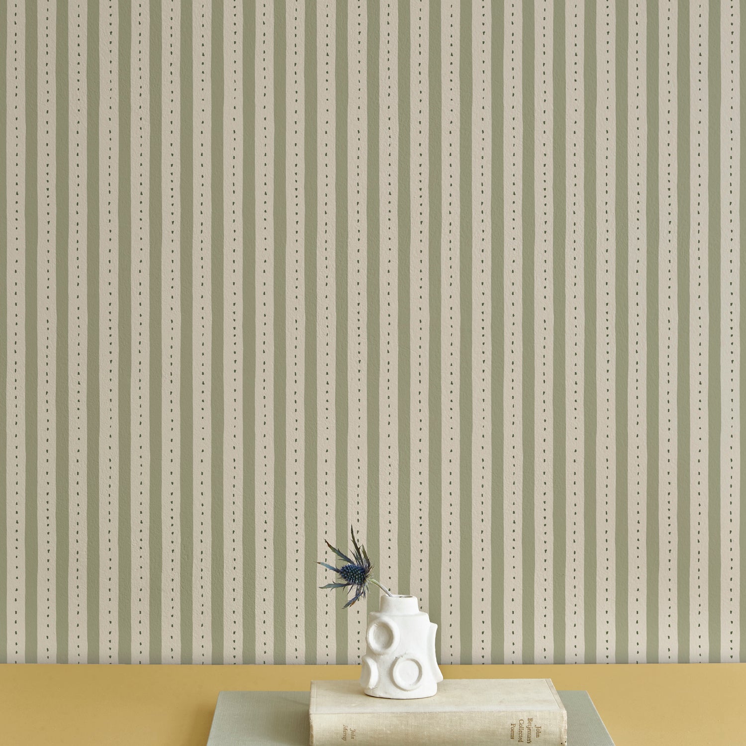 A stack of booked with a vase and thistle, against a wallpaper playful hand drawn dotted stripe in soft green.