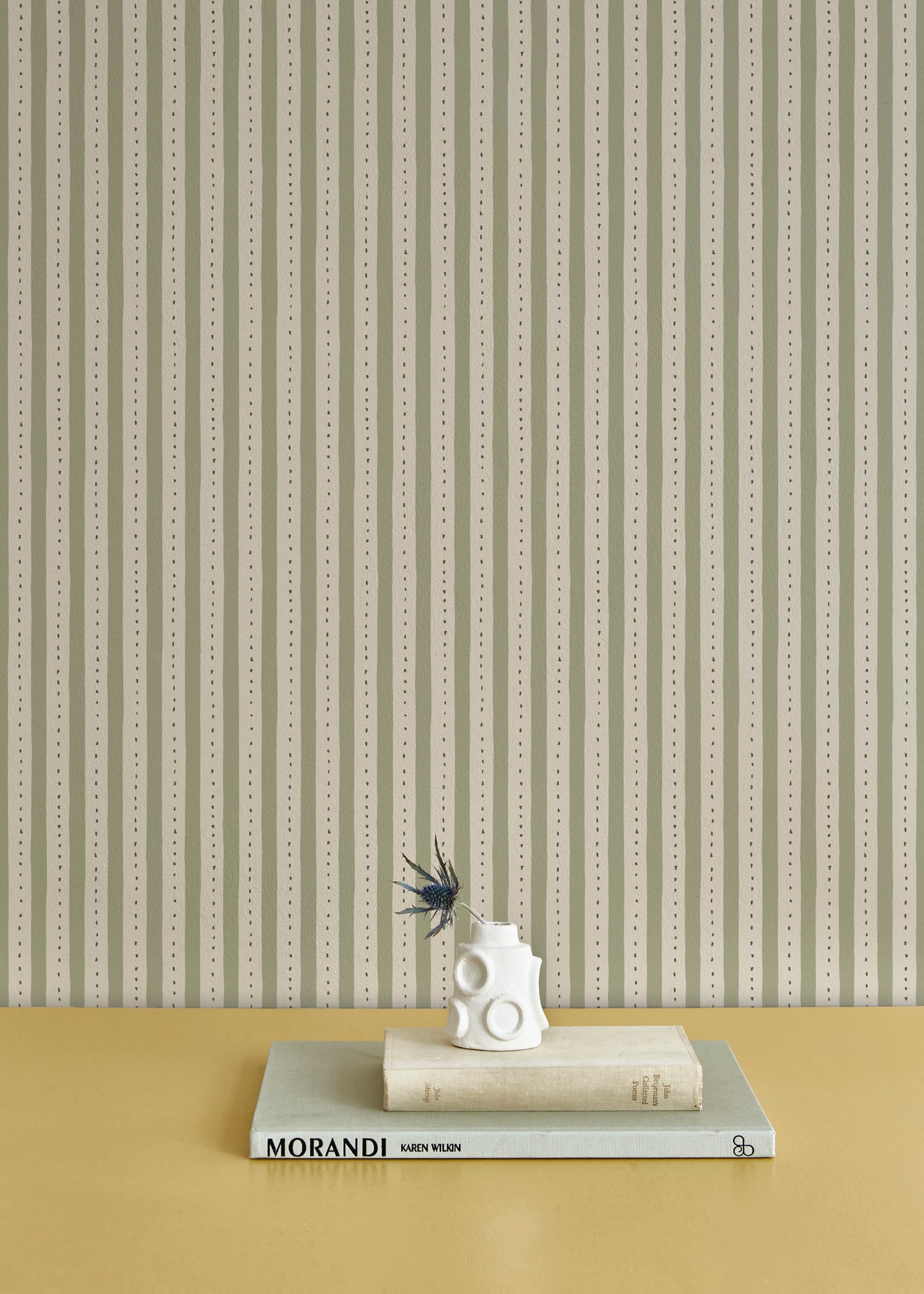 A stack of booked with a vase and thistle, against a wallpaper playful hand drawn dotted stripe in soft green.