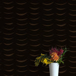 A vase of flowers stands in front of a wall papered in an abstract crescent print in brown on a black field.